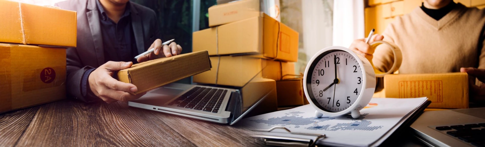 Personas preparando paquetes y documentos con reloj y laptop, gestión de pedidos y tiempos para evitar retrasos y sobrecostos.